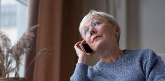 An elderly woman with short white hair is sitting by a window, holding a smartphone to her ear. She is wearing glasses and a gray sweater, and appears to be engaged in a phone conversation.